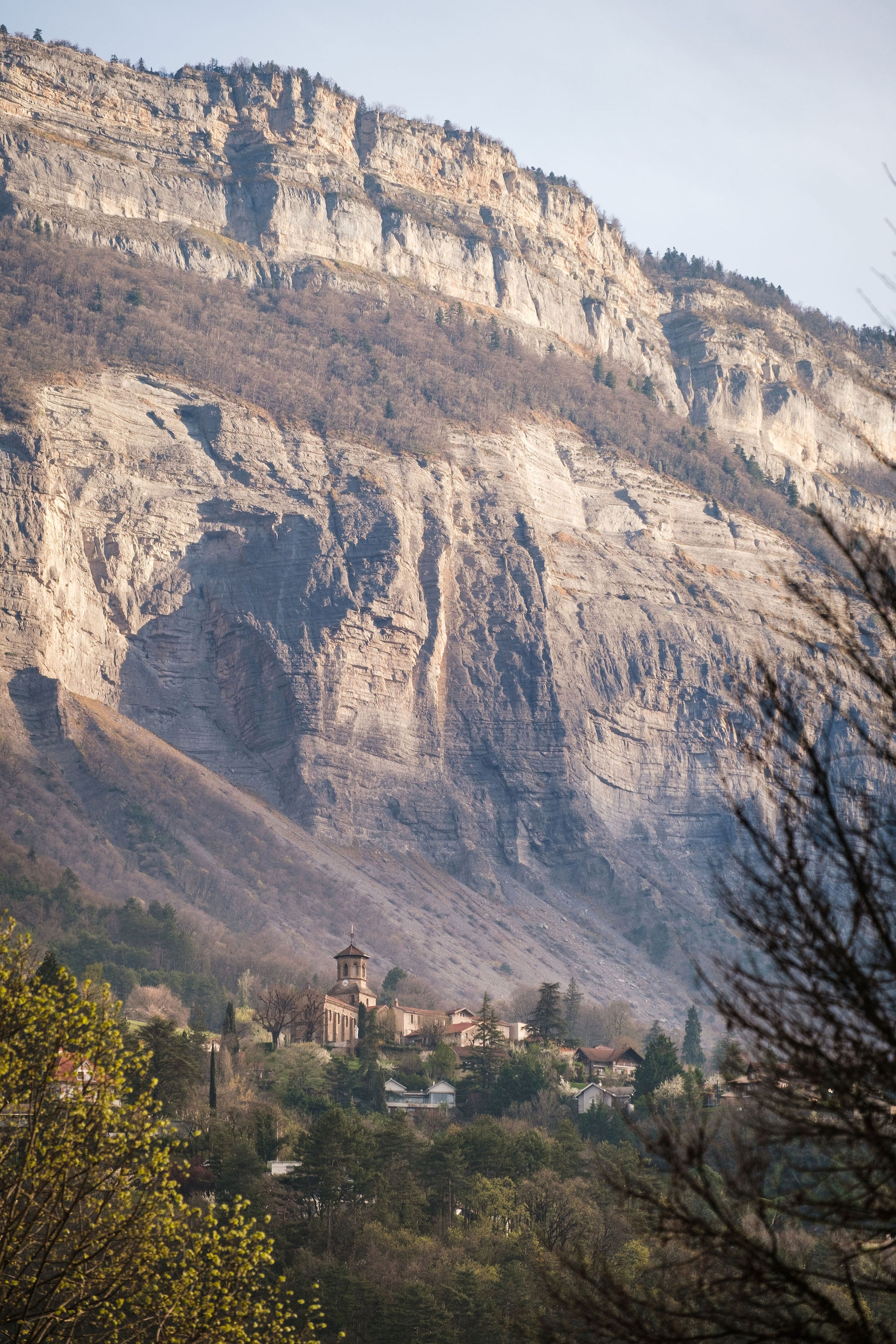 Small village with church at base of large rocky mountain with trees in foreground.