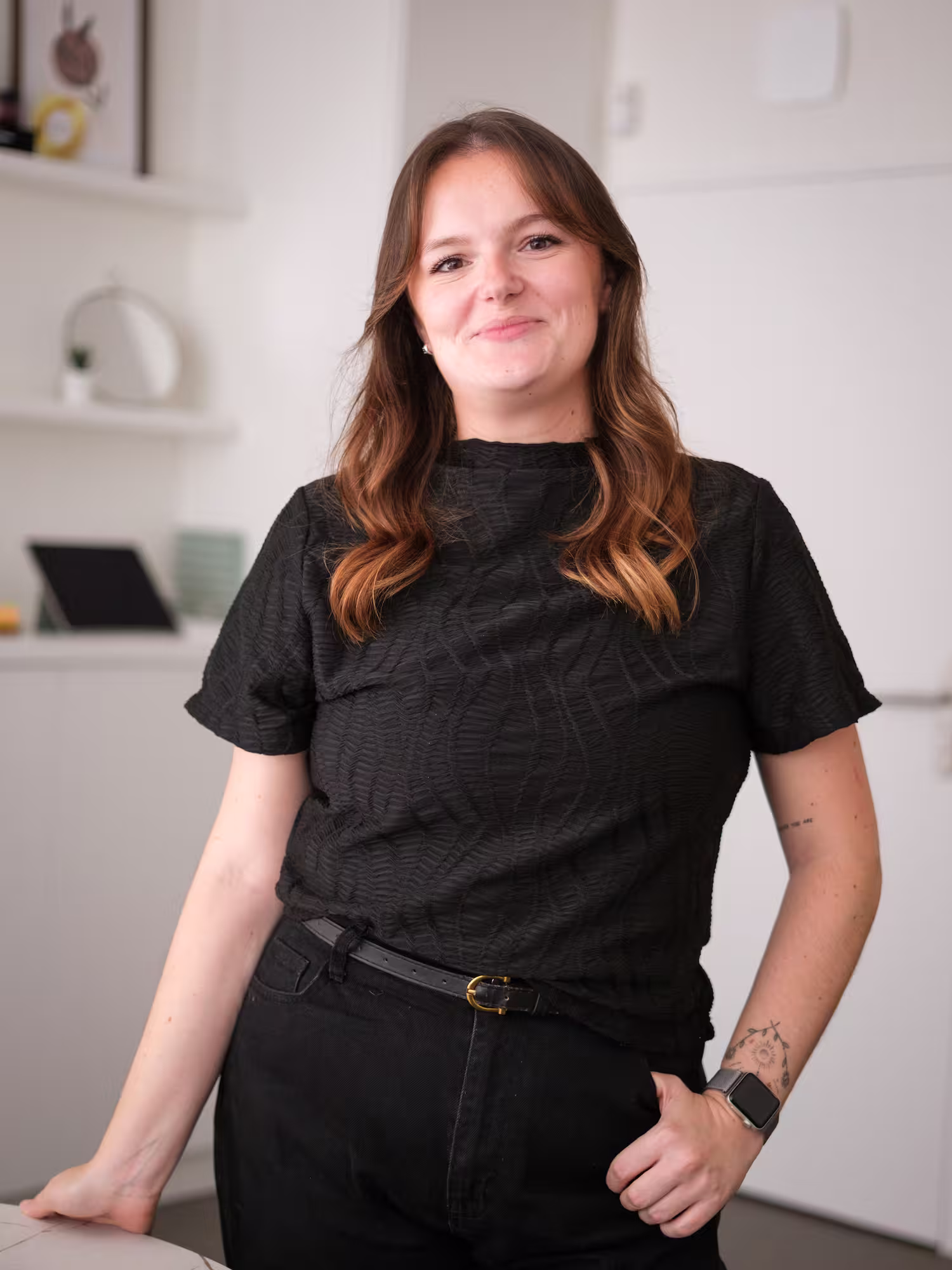 Smiling woman with curled brown hair wearing black lace sleeve top, in a softly lit room.