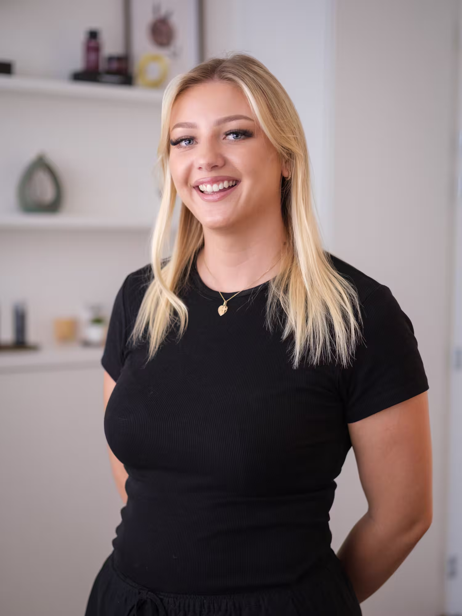 Smiling woman with blonde hair stands with arms crossed in a room with shelves and a mirror.