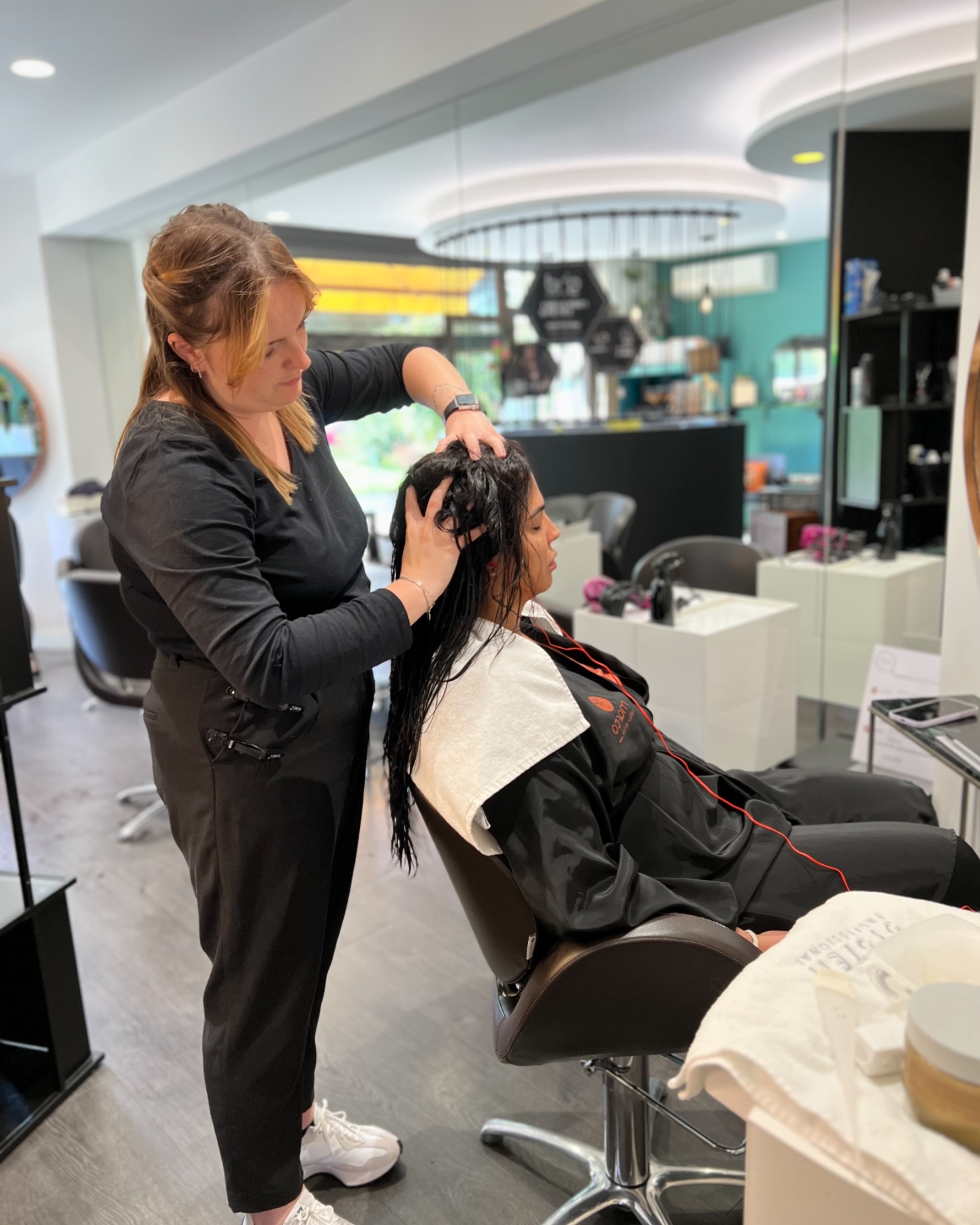 Hairdresser massaging wet hair of a seated woman in a modern salon with mirrors and chairs.