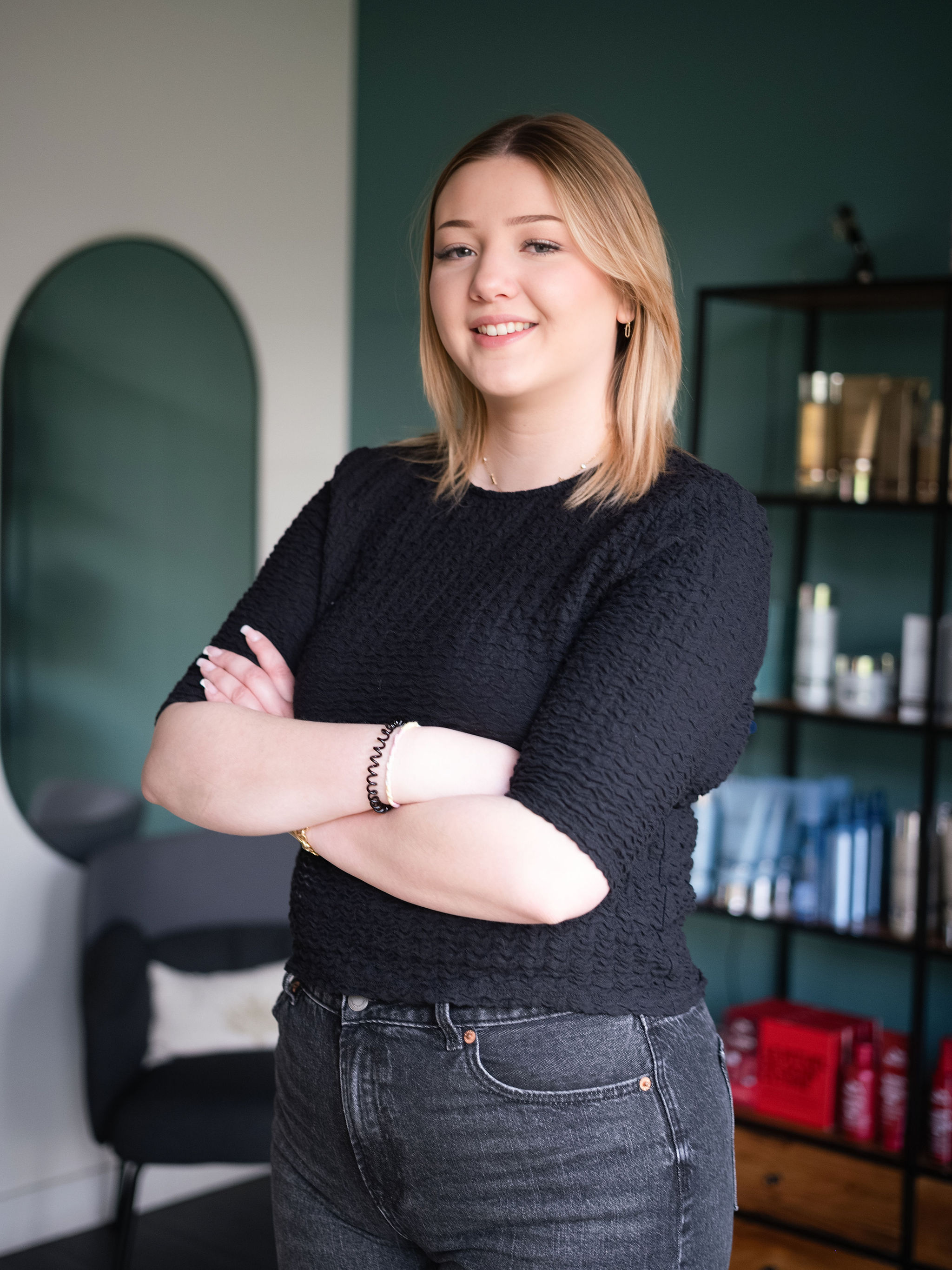Young woman with blonde hair in black top and jeans smiling with arms crossed in a room.