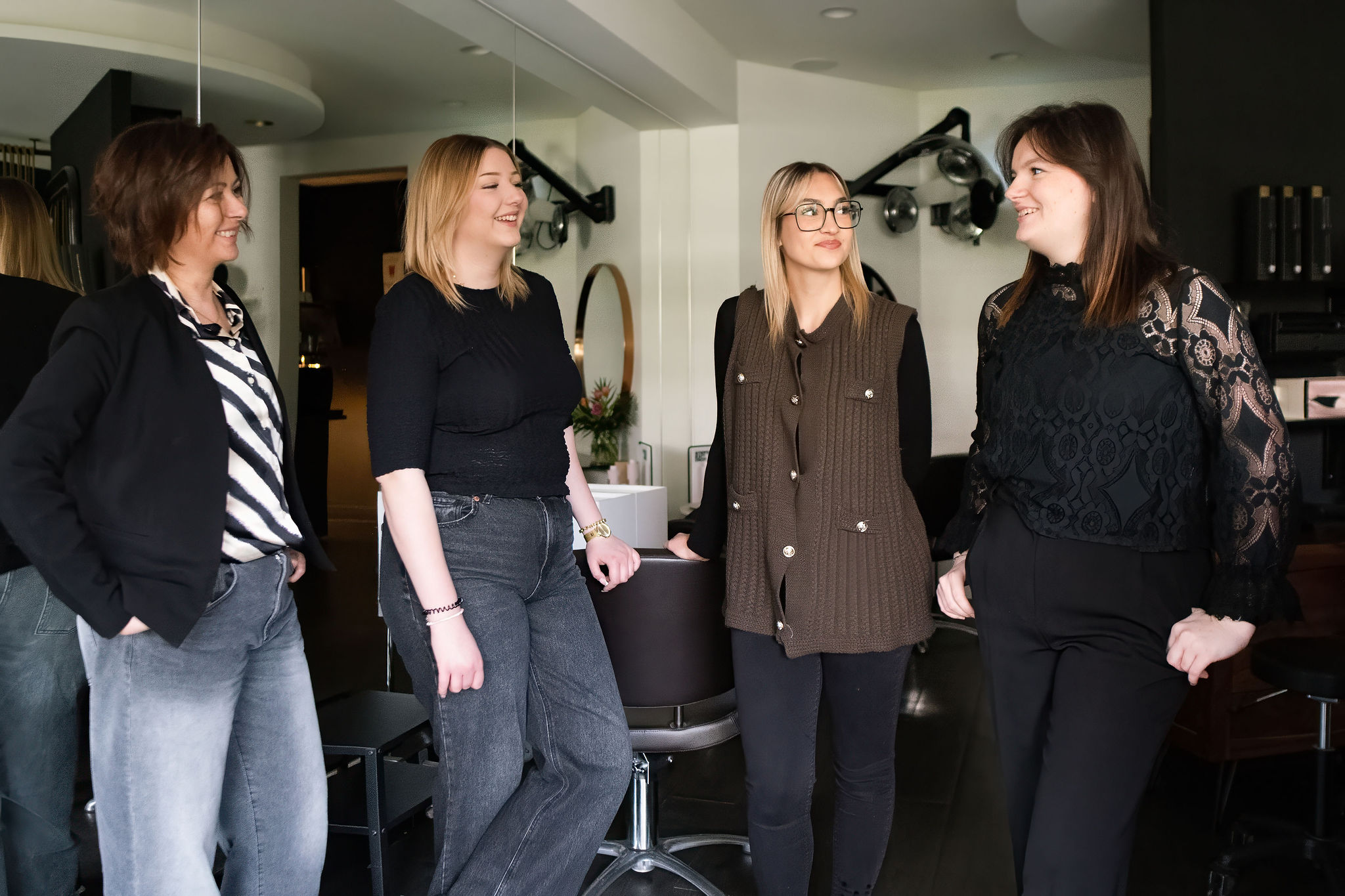 Four women standing and smiling indoors, casually dressed in black, gray, and brown clothes.
