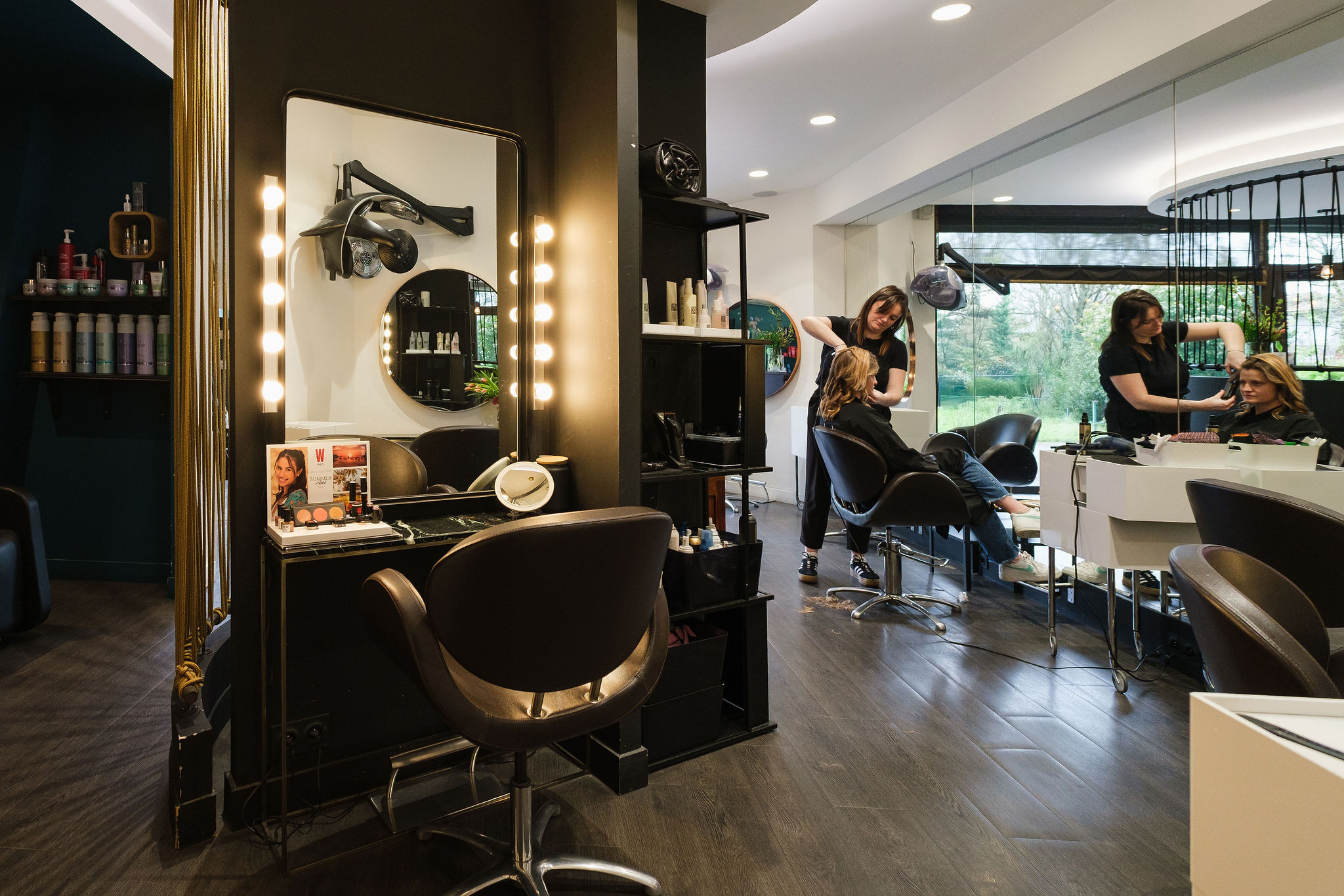 Hairdresser styling a seated woman's hair in a modern salon with lit mirrors and salon products.
