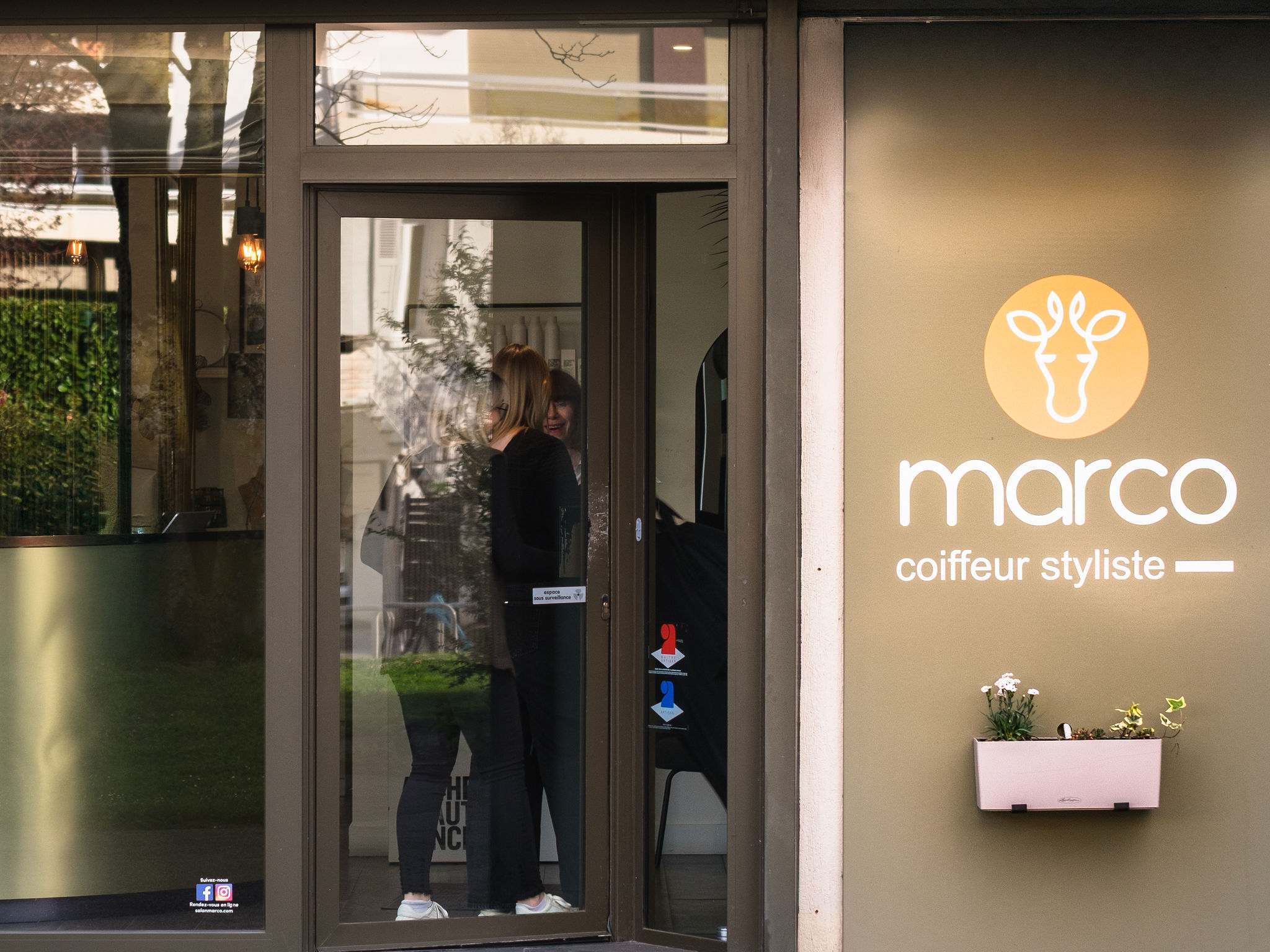 Glass door entrance of Marco coiffeur styliste salon with two people talking inside.