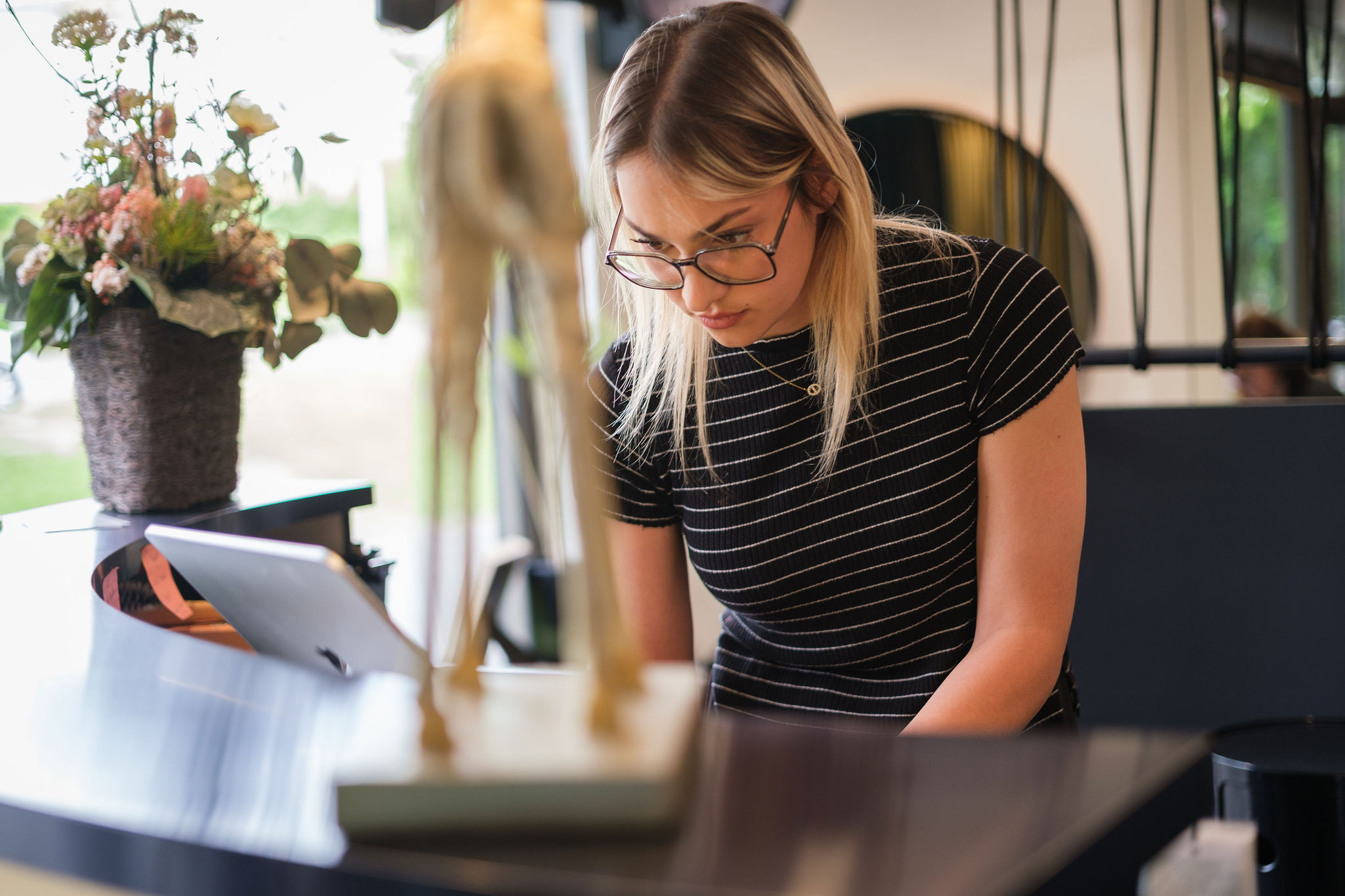 Young woman wearing glasses focused on a laptop at a café table with a flowerpot nearby.