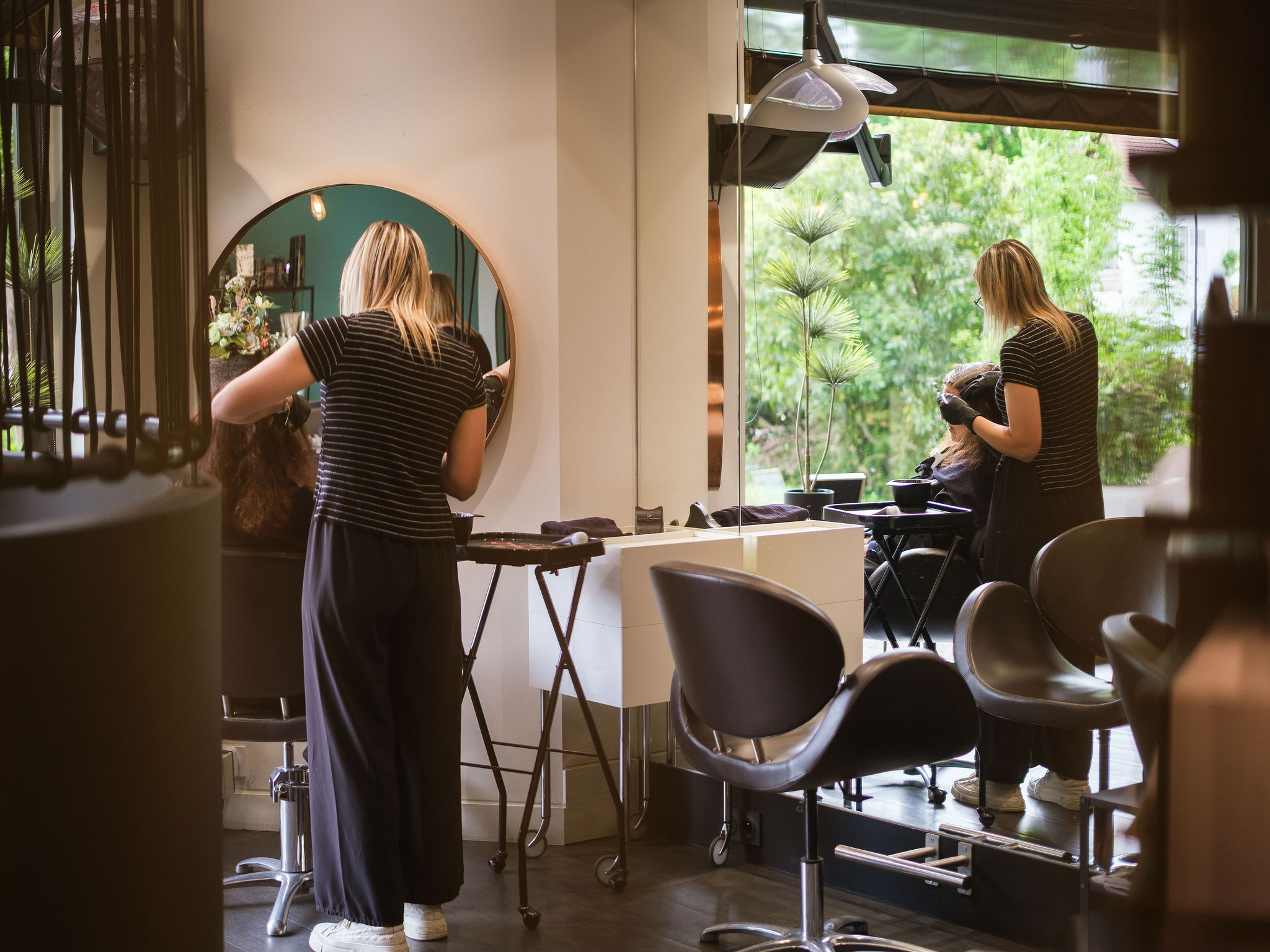 Hairdresser wearing gloves applying hair dye to woman seated in a salon chair by a large window.