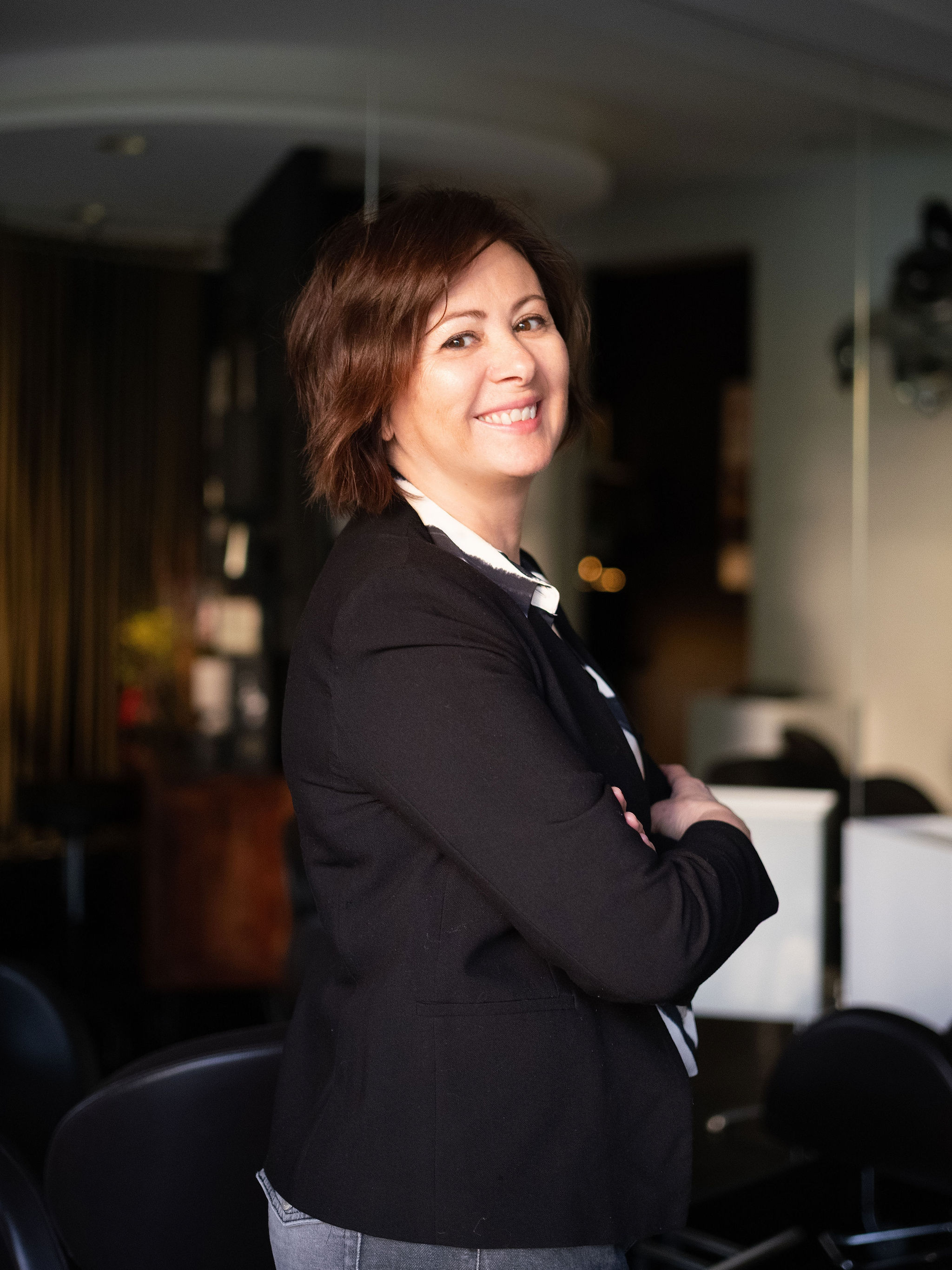 Smiling woman with short brown hair in a black jacket standing with arms crossed indoors.
