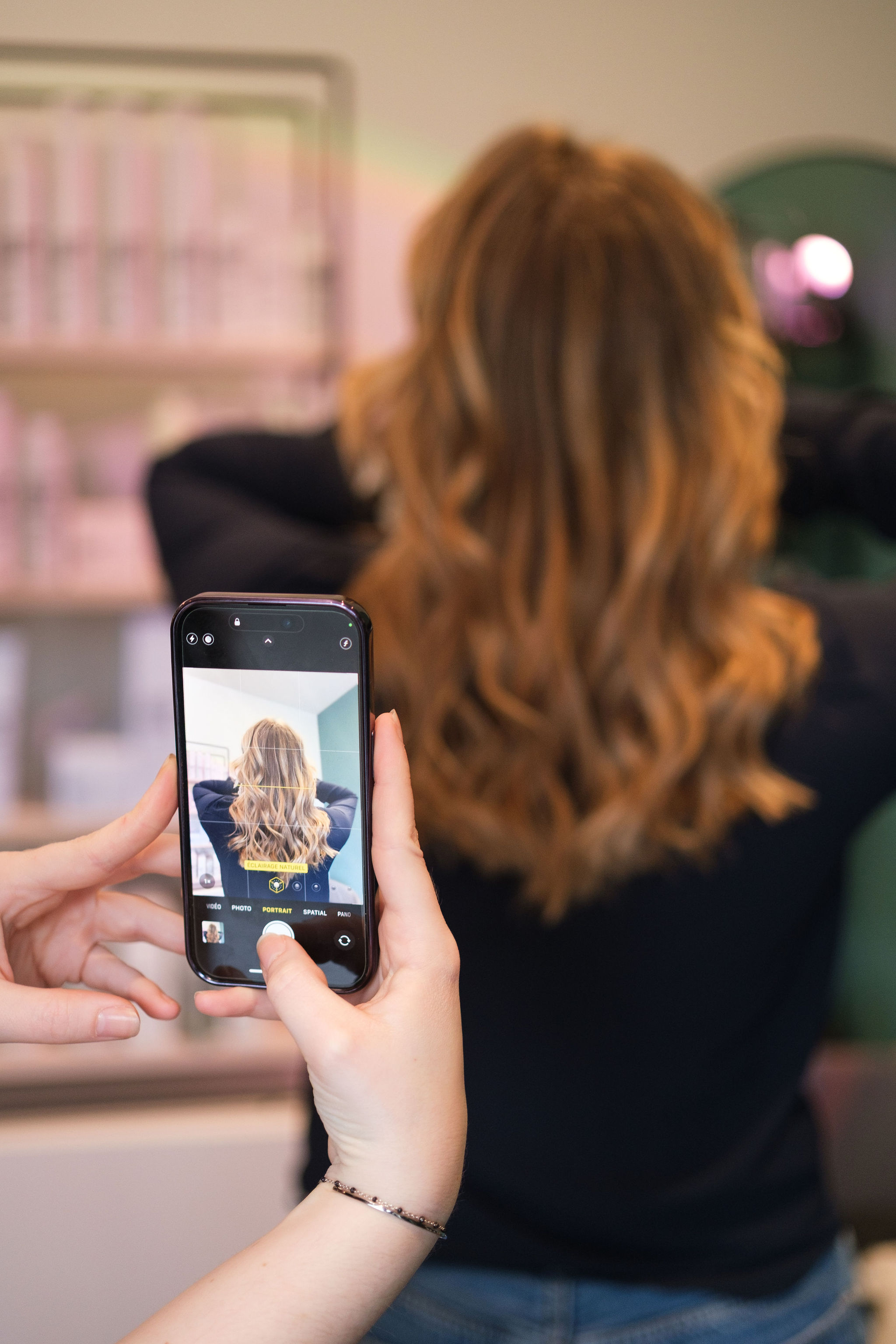 Person holding a phone taking a photo of a woman with long wavy hair in a salon setting.